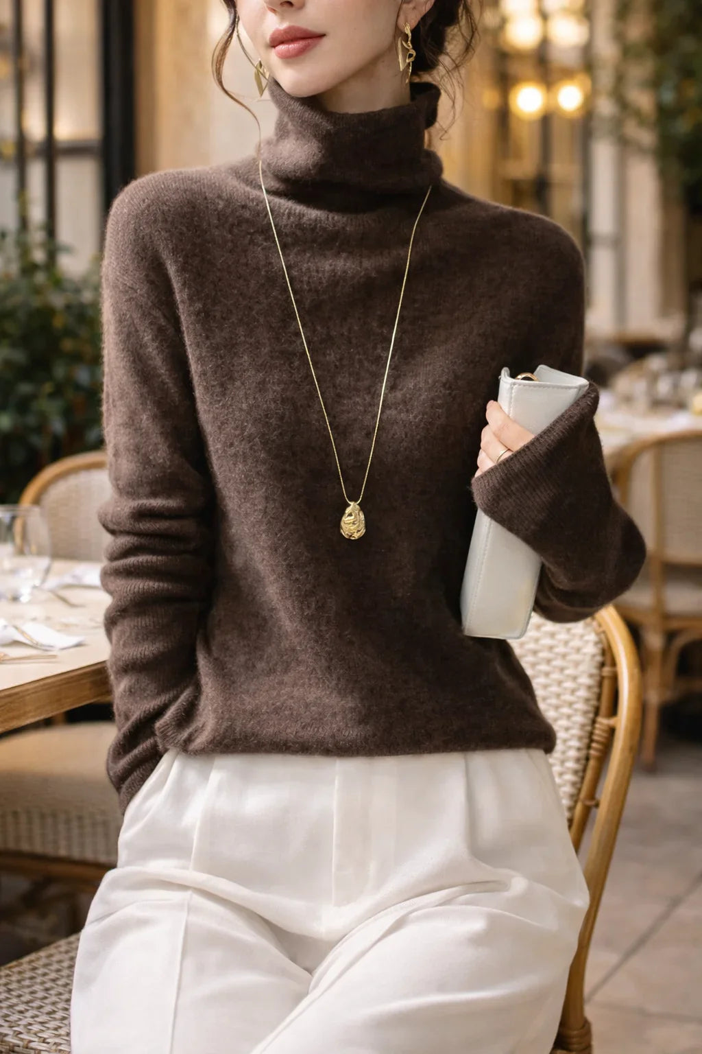 Femme élégante en pull col roulé marron, pantalon blanc, collier doré, tenant une pochette