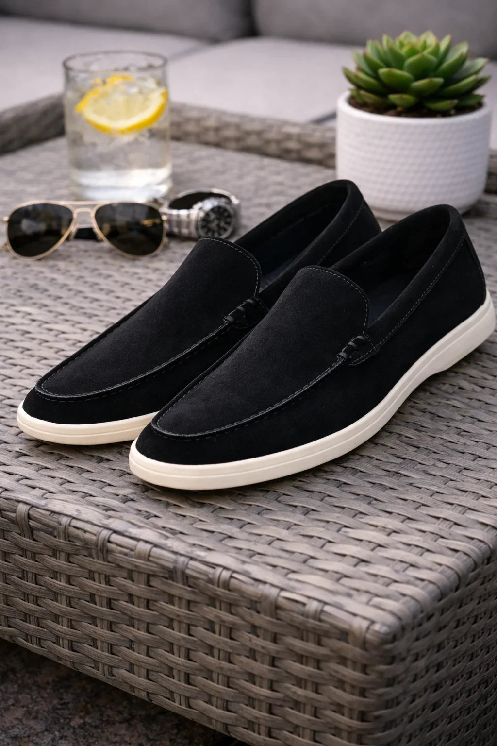 Mocassins noirs en daim pour homme à semelle blanche posés sur une table en rotin.